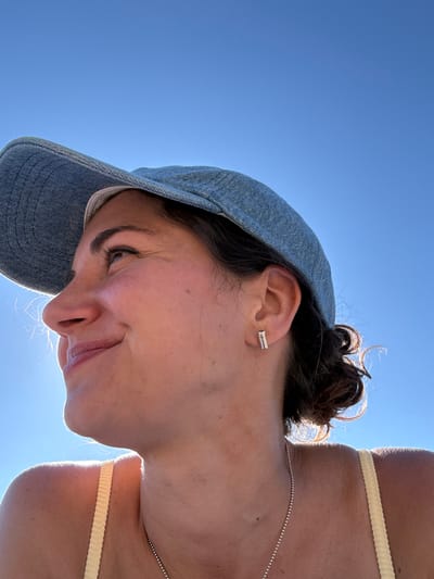 This is a close-up, low-angle outdoor photograph of a young woman wearing a denim baseball cap and a light yellow top against a clear blue sky. The sun is positioned behind her head, creating a lens flare that illuminates her hair and highlights her facial features. She is looking up and to her left with a subtle smile. She has a silver bar earring in her visible ear. The photograph captures her face from the nose upwards and a portion of her shoulder and neck. The overall impression is one of warmth and natural light. The image does not contain information about the location within the city of Рыбхоз, Russia.