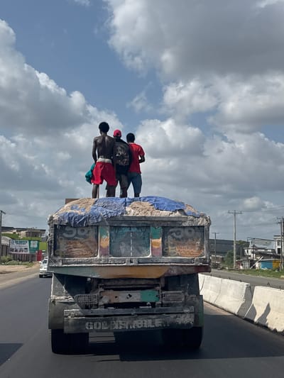 Three men are riding on top of a truck in Obinze, Nigeria. The truck bed is covered with a blue tarp and various markings. The men are dressed in casual attire, with one wearing a red shirt, another a black shirt, and the third wearing red shorts. The sky is partly cloudy. Buildings and power lines are visible in the background.