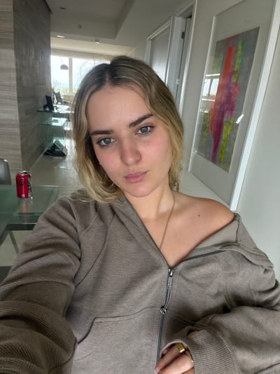 A fair-skinned individual with blonde hair and green eyes is facing the camera while wearing a beige hooded zip-up jacket and a gold ring. A gold necklace is visible. The subject is indoors, in a room with modern decor, including a glass table on which there is a crushed Coca-Cola can. A framed painting hangs on the wall. Outside the window, trees and blue water are visible. The location is likely Key Biscayne, United States of America.
