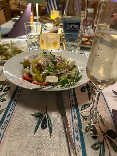 The scene depicts a dining table at Restaurant Rhodos in Eppelheim, Germany. In the immediate foreground on the right, a tall, clear champagne flute containing a sparkling, pale yellow beverage with condensation and visible bubbles is positioned. To its left, a white circular plate holds a Greek salad, featuring diced cucumbers and tomatoes, sliced red onions, green peperoncini, black olives, and cubed feta cheese, garnished with fresh herbs. A silver-colored knife with a dark handle is placed horizontally to the left of the salad plate. Both the knife and the salad plate rest on a light-colored placemat or table runner patterned with dark green olive branches and blue decorative stripes.

Behind the salad, a clear glass tumbler holds water, and further back, a blue-labeled bottle of "Seltzers Classic" mineral water stands within another clear glass. Between these, a small, illuminated candle holder with a subtle pattern is visible. A portion of a second white plate is present on the far left, containing green peppers and a white topping. The background is softly blurred, revealing a restaurant interior with other tables draped in white tablecloths, chairs, and diffuse warm lighting. A miniature Greek flag on a pole is identifiable behind the mineral water bottle, and additional dishes, including bread slices, are discernible on adjacent tables. No individuals are visible.