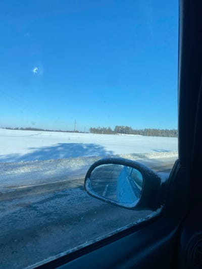 The image depicts a winter landscape in Пліскі сельскі Савет, Belarus, viewed from the interior of a vehicle. The perspective is through a passenger-side window, with the vehicle's dark door frame and partially snow-encrusted side mirror visible on the right. Outside the vehicle, a dark, partially cleared road or shoulder runs horizontally across the foreground, showing icy or wet patches and residual snow. Beyond the road, a vast, flat field is completely covered in pristine white snow. Long, dark shadows are cast across the snow-covered field, extending from the left side of the frame. In the mid-distance, a series of utility poles with power lines traverse the landscape. A dense line of leafless deciduous trees and some coniferous trees forms the distant tree line against the horizon. The sky is uniformly clear and bright blue, indicating a sunny day. A distinct bright flare or imperfection is visible on the window in the upper left quadrant of the image. No people are visible.