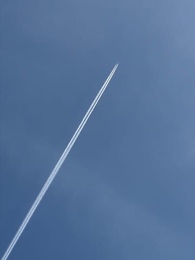 The image shows a contrail in a blue sky. The contrail is a white line stretching diagonally across the frame from the lower left to the upper right. The contrail appears to be produced by a jet plane, which is not visible. The sky is a solid blue color with slight variations in tone. There are no other objects or people visible in the image. The image is not in the city of Ol'khovo, Russia.