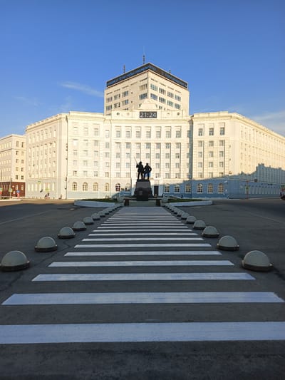 In Norilsk, Russia, a wide pedestrian crossing with white stripes is in the foreground. Beyond the crossing, a plaza features a monument with two standing figures. Behind the monument is a large, imposing white building with many windows, featuring a digital clock displaying "21:24" on its facade. The building's upper section has a blue banner with white text. The sky is clear and blue. The overall scene is set in an urban environment under daylight.