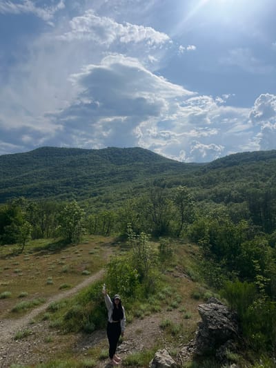 The image shows a person standing on a dirt path in a grassy area in Kamchia, Bulgaria. The person is wearing dark clothing with a light-colored jacket, a hat and holding a plastic bottle. Behind the path is a steep hill covered in dense green forest. Above and to the left, there are puffy white clouds in a light blue sky, with bright sunlight shining from the top left. The image appears to have been rotated to the right.