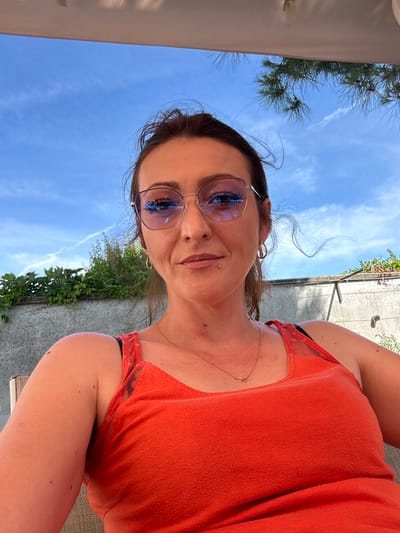 A woman is taking a selfie outdoors under a blue sky. She is wearing an orange tank top and eyeglasses. The background consists of a wall, greenery, and a tree branch. The woman has brown hair and appears to be in her 30s or 40s. She is smiling and looking at the camera. There is no indication of the specific location within Villa Pasquali, Italy.