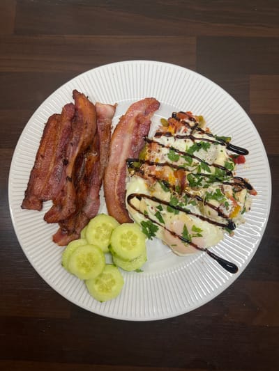 A white plate containing a breakfast of fried eggs, bacon, and sliced cucumbers is displayed on a dark wood table. The fried eggs are topped with chopped vegetables and herbs, and drizzled with a dark sauce. The bacon is cooked to a crisp and arranged to the left of the eggs. Several slices of cucumber are placed to the bottom left of the plate. The table surface suggests a domestic setting. There are no individuals present. The image provides a clear, overhead view of the meal.