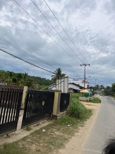 The image depicts a street scene in Rantau Pauh, Indonesia. In the foreground, a black metal gate with gold accents is partially open, revealing a paved area behind it. The gate is bordered by a beige wall. To the right of the gate is a patch of overgrown grass and dirt, with some scattered litter, including a crumpled white item and a blue object.

Beyond the gate, a paved road curves to the right, with a dirt and grass shoulder. A series of power lines with wooden poles are visible overhead. Along the road, there are several buildings. One building is white with a red roof and appears to be a house or a small commercial establishment. Another building has a white facade and a light-colored roof, with an attached yellow awning supported by poles, suggesting a small outdoor stall or market. Further down the road, more buildings are visible, some with lighter colored roofs, and a few small trees and palm trees dot the landscape.

In the background, a lush green hillside is covered with dense vegetation, including palm trees and other tropical foliage. The sky is overcast with heavy gray clouds, indicating a cloudy day. The overall impression is a rural or semi-rural setting in Indonesia.