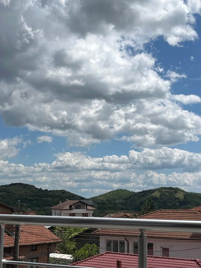 The image depicts a view from a balcony in what appears to be Kornitsa, Bulgaria, evidenced by the architecture and landscape. The foreground features a metal railing, partially obscuring a row of houses with red tile roofs and white facades. Behind the buildings rises a green, undulating hillside. The sky is filled with a mixture of cumulus and stratocumulus clouds, creating a textured pattern against patches of blue. The image is shot during daylight with diffused lighting.