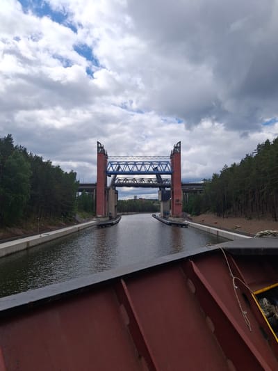This image captures a view looking forward along the Södertälje Canal in Södertälje, Sweden, from the foredeck of a vessel. The central feature is the Södertälje swing bridge, characterized by two prominent, red-painted concrete towers that flank the canal. A blue steel truss structure, likely the movable section of the bridge, spans between the tops of these towers. Beneath this main structure, a lower, darker blue or grey truss element is visible.

Behind the swing bridge, two additional bridges cross the canal. The closest is a multi-lane concrete highway bridge, identified as the E4/E20 Södertälje highway bridge, visible at a higher elevation. Further in the distance, a darker, older-looking bridge, likely the railway bridge, is faintly discernable.

Both sides of the canal are lined with dense coniferous forest, primarily pine trees. On the left bank (viewer's left), a concrete pathway or service road runs parallel to the canal, equipped with several short light posts. On the right bank, the forest extends to the canal edge, with some areas of disturbed earth or sand visible between the water and the trees. In the far distance, beyond the bridges and forests, urban structures including buildings and a water tower are visible on the horizon, confirming a city setting.

The foreground of the image shows the rust-red painted steel plating of the vessel's hull or deck structure, with internal support beams visible on the right. A dark, flat beam or railing runs horizontally across the top edge of this structure. On the far right, a coiled rope, likely a mooring line, is visible.

The sky is heavily overcast with a mix of large, grey clouds and patches of brighter blue sky breaking through in the upper left quadrant. The lighting suggests daytime conditions. No people are visible in the scene.