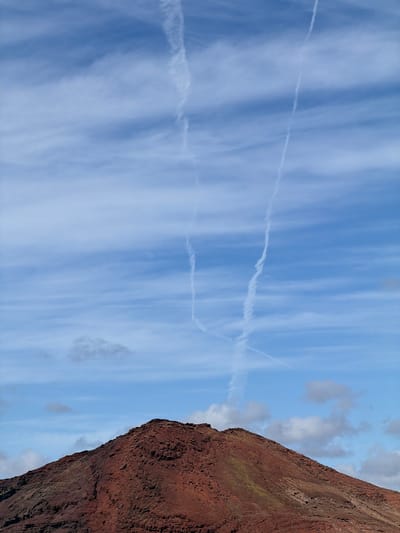 The image shows a scene in Yaiza, Spain, featuring a reddish-brown volcanic rock formation on the right side of the frame contrasting with a blue sky filled with white clouds and contrails in the background. The contrails stretch across the sky, and the clouds appear streaky. The rock formation appears textured and layered.