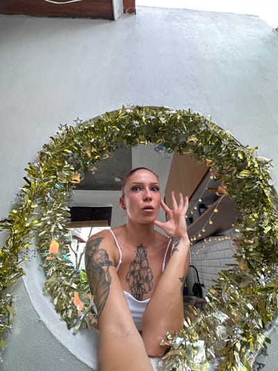 The scene, located in Kerobokan, Indonesia, centers on an individual, appearing female, positioned within a large circular opening or mirror. This circular feature is elaborately decorated with a dense, shimmering gold and silver tinsel garland, interspersed with small silver star cutouts and iridescent bell-shaped ornaments. The individual has light-toned skin, light blue eyes, and pink-toned hair pulled back. She exhibits a small nose ring on her left nostril and a stud on her right earlobe. Prominent tattoos include a large floral design on her upper chest/sternum, an intricate pattern on her left upper arm, and a rose-like design on her left forearm. She wears a white tank top. Her expression is characterized by wide eyes and a slightly open mouth, while her right hand is raised near her face with fingers loosely spread. The background, visible through and reflected in the circular element, includes: on the upper right, wooden cabinetry with a string of warm-toned spherical lights hanging below and a white subway tile backsplash; on the lower left, an outdoor view with a tree, a visible bamboo blind, and a portion of a light-colored building featuring a dark railing, consistent with local architecture.