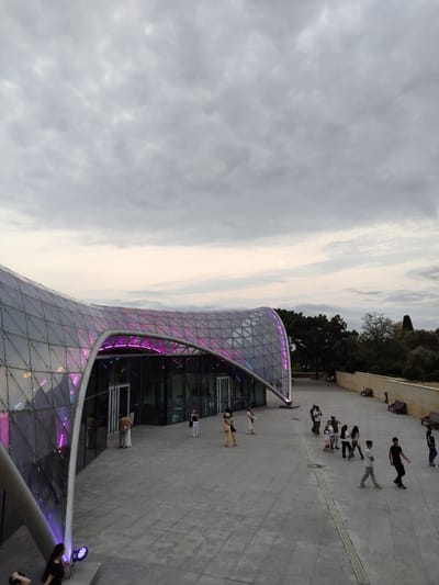The image captures an outdoor view of the Heydar Aliyev Center in Sabail Raion, Azerbaijan, under a cloudy sky. The architectural centerpiece is a modern building with a sweeping, curvilinear roof made of glass and a metallic grid structure, illuminated with purple lights along its edge. Several people are walking on the paved plaza in front of the building, some individually and some in small groups. The plaza is expansive, with a drainage grate running horizontally across it. In the background, trees and a low wall with benches are visible. A faint sun is discernible through the clouds. The overall scene suggests a public space utilized for pedestrian movement and possibly as a tourist attraction.
