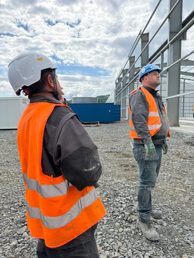 Two construction workers are standing on a gravel-covered construction site in Wilkowisko, Poland. The worker in the foreground, on the left, is wearing a white hard hat and an orange high-visibility vest over a dark gray work jacket. They are seen from the back and are looking upwards. The worker on the right is wearing a blue hard hat and an orange high-visibility vest over a dark gray jacket, blue jeans, and work boots. They are also looking upwards. In the background, a large steel structure of a building under construction is visible, along with shipping containers and other construction equipment. The sky is cloudy.