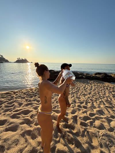 Two individuals are on a sandy beach in Castell d'Aro, Platja d'Aro i s'Agaró, Spain, during sunrise. The person on the left, a woman with her hair in a bun and a tattoo on her ribcage, is wearing a bikini bottom. She is holding a large plastic bottle of water, offering it to the person on the right. The person on the right, wearing a baseball cap and a bikini top, is reaching out to take the bottle. The sun is low in the sky, casting a warm glow on the water and the beach. The ocean is calm with gentle waves, and rocks are visible along the shoreline. A small island with trees is in the background. The sky is clear and blue, transitioning to a lighter shade near the horizon.