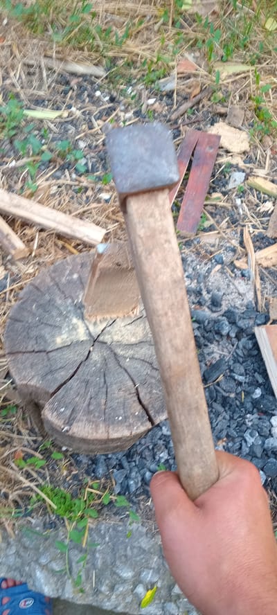 The image shows a person's hand holding a wooden-handled tool, possibly an ax or a splitting maul, positioned above a cut log. The tool's head appears to be made of metal and has a square shape. The log is resting on the ground, which is covered with dry grass, twigs, and some dark material that looks like charcoal or small rocks. There are also small green plants growing amongst the debris. The scene suggests an outdoor setting, likely in a rural area, given the presence of a log and natural ground cover. The lighting appears to be natural, suggesting it is daytime. No specific weather conditions are evident, but the dry grass might indicate a dry period. The image does not convey any discernible emotions, as the focus is on the action and the objects. There is no visible text in the image. The location context provided is Iskrets, Bulgaria.