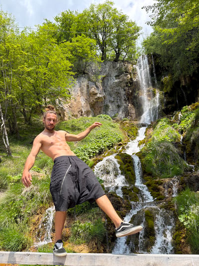 A man stands shirtless in front of a waterfall in the Prijepolje Municipality, Serbia. He wears black shorts and black-and-white sneakers. He stands on a wooden structure, likely a viewing platform, with his arms outstretched. The waterfall cascades over mossy rocks into a pool below, surrounded by lush green vegetation and trees. A rock face is visible behind the waterfall. The sky appears overcast.