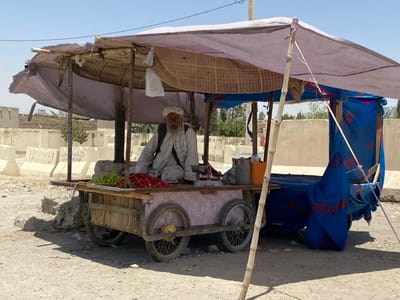 The image shows a man sitting in a makeshift stall, possibly a fruit stand, on a dirt road in Dand, Afghanistan. The stall is constructed from a wooden cart with a canopy made of a large tarp. The man is wearing a long white robe and a white cap. He has a long white beard. There are two large, metal wheels on the cart and a smaller metal wheel on the back. The stall appears to be located in a semi-urban area with concrete structures and dirt roads. The man is selling fruit, including green apples and red tomatoes, from a wooden crate on the cart. He appears to be waiting for customers. The image was taken in the afternoon with a blue sky overhead. There is a large blue tarp on the right side of the stall and a small tree behind the stall. The man is sitting on the right side of the cart, next to a large water jug and a red bucket. The image was likely taken on a sunny day, with no clouds in the sky. The image has a resolution of 1080 x 1920 pixels. There are some buildings behind the cart, suggesting that the stall is situated within a town or city.