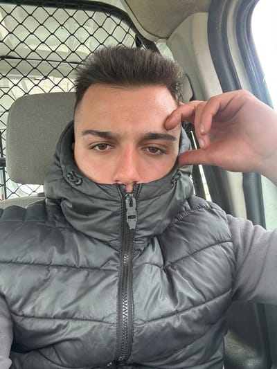 A young adult male is depicted seated inside a vehicle, located in the city of Srednia, Bulgaria. He has short, dark hair styled upwards and brown eyes, looking directly at the viewer. His face is partially obscured by a dark grey hooded puffer vest, which is fully zipped up to just below his nose. He is also wearing a grey long-sleeved garment under the vest. His left hand is positioned with the thumb and forefinger touching his forehead. Behind him, a grey fabric headrest and a black metal mesh partition are visible, consistent with a commercial vehicle interior. The background beyond the vehicle's interior is indistinct but appears to be an outdoor environment under overcast conditions.