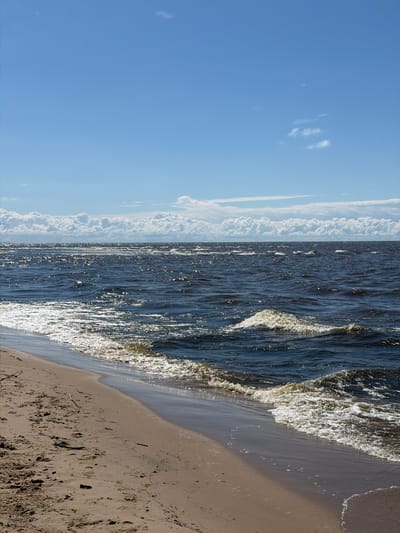 This is a vertical, eye-level, medium shot of a sandy beach meeting a dark, choppy sea under a blue sky with scattered white clouds. The sand is light brown with some small pieces of debris, including twigs and seaweed, and is marked with footprints. The sea is a deep blue, with white foam and cresting waves visible along the shore. Sunlight glints off the surface of the water, creating a shimmering effect. In the distance, along the horizon, there are more white clouds forming a band across the sky. The image captures a natural coastal environment. The location is the beach in Gauja, Latvia. There are no people visible in the image.