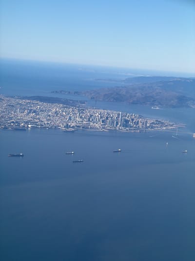 An aerial photograph captures a panoramic view of a major coastal city and its expansive bay under a clear, bright sky. The image, best viewed rotated clockwise by 90 degrees, places a densely developed urban area along the bottom and left side, stretching out into a vast body of water on the right.
The cityscape is characterized by numerous buildings, forming a skyline that indicates a bustling metropolitan center. In the mid-distance on the left, the iconic red-orange towers of the Golden Gate Bridge are visible, spanning a strait and connecting the main city to a hilly, mountainous landmass. This landmass, likely the Marin Headlands, is covered in natural terrain.
The wide body of water, indicative of a bay or estuary, is a deep blue and appears calm. Several large cargo ships or tankers are scattered across the water, either anchored or in transit, with a few smaller vessels also discernible by their faint wakes. The bright, even lighting suggests it is daytime, likely morning or afternoon, with excellent visibility. No people or legible text are visible due to the high altitude. The scene portrays a significant urban environment with substantial maritime activity, strongly identifying the location as San Francisco Bay, consistent with the provided Oakland context.