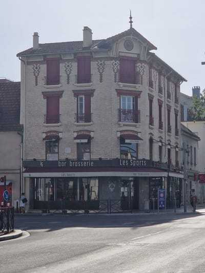 The image shows a three-story building with a facade of light-colored bricks and red trim, located on a street in Antony, France. The building has a mansard roof with dormer windows and chimneys. The ground floor features a commercial establishment with a sign that reads "Les Sports" and "bar brasserie." Large windows with dark frames look out onto the street. The street in front of the building is paved and shows some sunlight reflections.

On the sidewalk, a woman in a light-colored dress and a dark handbag is standing to the left of the entrance, facing the building. Further to the right, a person wearing a red top and dark pants is standing near the entrance of another building. A metal fence runs along the sidewalk in front of "Les Sports." Various street signs are visible, including a speed limit sign (20) and a sign indicating priority for pedestrians. The sky is clear and bright, suggesting daytime.