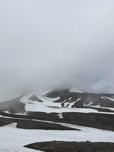 The image shows a desolate, mountainous landscape under a heavily overcast sky. The foreground consists of dark, volcanic-like earth interspersed with patches of snow and ice. The midground reveals undulating hills covered with patches of snow and dark earth, leading to steeper slopes. The background is dominated by a large mountain, its peak obscured by dense fog or clouds. Streaks of snow define the contours of the mountain. The scene appears to be a natural, undeveloped area, possibly within or on the outskirts of Petropavlovsk-Kamchatskiy, Russia, given its volcanic terrain and cold climate. No people or specific actions are visible. The environment is characterized by rugged terrain, snow cover, and limited visibility due to the weather.