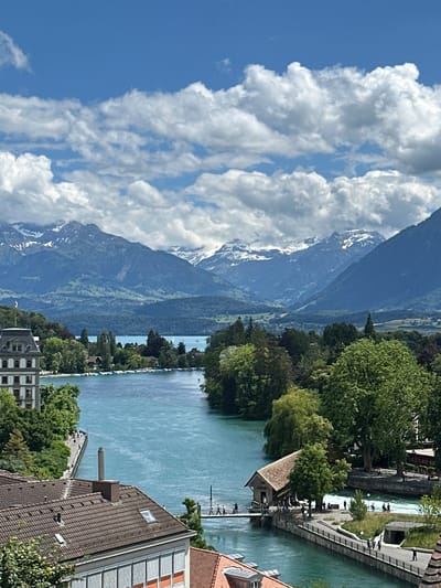 This is an outdoor, high-angle, eye-level shot of a river flowing through the city of Thun, Switzerland, with mountains in the background. The river is a vibrant turquoise color and is surrounded by lush green trees. On the left side of the river is a large, old building with a red-tiled roof and white window frames. Further along the river, there is a bridge with several people walking across it. In the background, snow-capped mountains rise majestically against a blue sky dotted with white clouds. The scene is peaceful and picturesque, capturing the natural beauty of Thun and its surroundings.