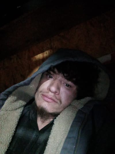 The scene is identified as being in Ozark, United States. A single male individual, appearing to be a young adult, is depicted in a close-up, dimly lit environment. He possesses dark, curly hair and dark facial hair consisting of a mustache and a goatee. The subject is wearing a dark, hooded jacket with a light-colored, possibly sherpa-like, lining visible around the hood and collar, worn over a dark, patterned shirt. A strong, singular light source illuminates the left side of his face (viewer's right), casting the right side into deep shadow. His eyes are open and directed towards the viewer, with a neutral expression. The background consists of dark, textured surfaces resembling unfinished wood paneling, such as oriented strand board, indicating an indoor, possibly enclosed or utilitarian space. No other individuals or distinguishing objects are visible.