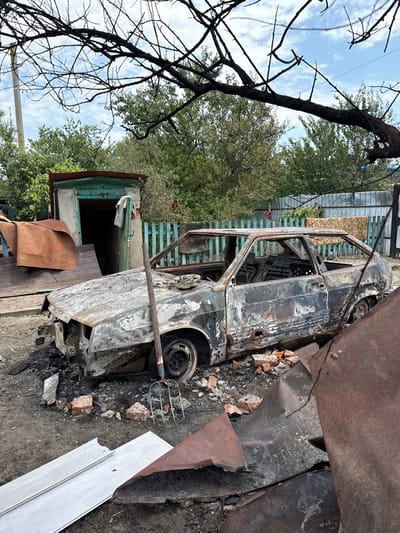 This image captures a static outdoor scene in Nedryhailiv, Ukraine, depicting the aftermath of significant damage. The central subject is the charred and extensively burned chassis of a grey two-door hatchback, consistent with a Lada Samara variant. The vehicle is situated on dark, uneven dirt ground, its exterior heavily discolored by fire, windows missing, and interior completely incinerated. Visible perforations, consistent with projectile impacts, are present on the body panels, particularly on the driver's side. The front bumper is absent, and the engine bay appears gutted. A single black wheel remains on the front passenger side.

A wooden-handled pitchfork leans against the front left fender of the vehicle. The ground around the car is littered with debris including fragments of red brick, charred material, and twisted, rusted metal sheets. In the immediate foreground, large pieces of white paneling and additional twisted metal are visible.

In the background, a light blue wooden slat fence extends horizontally behind the vehicle. Further back, dense green trees and shrubs form a natural backdrop. To the far left, a small, square building with a dark green door and roof trim is partially visible, with folded brown fabrics or tarpaulins stacked nearby. A large, dark, bare tree branch extends into the upper portion of the frame, silhouetted against a partly cloudy blue sky. No individuals are present in the scene.