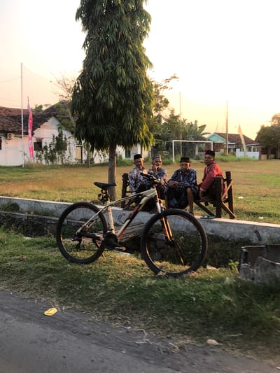 In Sentul, Indonesia, four elderly men wearing traditional Islamic skullcaps and patterned sarongs are seated on a wooden bench, conversing. In the foreground, a mountain bike is parked with its front wheel turned to the left. The scene is set in an outdoor area with a large tree to the left of the men, a building with a red-tiled roof in the background, and a sports field with a net and goalposts further back. The sky is clear and illuminated by the warm glow of what appears to be sunset. The ground is covered with grass and a dirt path.