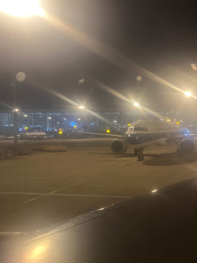This media file captures a nighttime scene at an airport, likely in Guangzhou, China. The primary subject is a large passenger airplane parked on the tarmac, viewed from a slightly elevated position, possibly from another aircraft or a building overlooking the scene. The airplane is predominantly white with visible engines.
The setting is an airport at night, characterized by numerous bright lights illuminating the tarmac and the airport terminal in the background. The terminal appears to be a modern structure with large glass windows reflecting the lights. Several smaller vehicles and airport equipment are present on the ground near the airplane, suggesting active ground operations.
The time of day is clearly night, with a dark sky and artificial lighting dominating the scene. The weather appears to be clear, as there is no visible precipitation or fog obscuring the view, although there might be some atmospheric haze or light bloom from the strong lights.
No people are explicitly visible, but the presence of an airplane and airport infrastructure implies human activity related to air travel. The overall mood conveyed is one of a bustling but serene nighttime airport environment.
Visible text includes yellow signs on the tarmac with what appear to be numbers, possibly gate numbers or directional information, but they are too small and blurry to be clearly read. The image is oriented vertically.