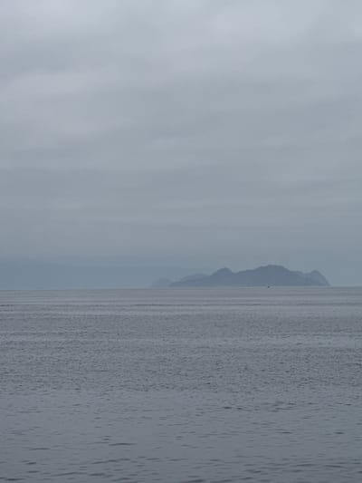 The image depicts a wide expanse of calm, gray water under an overcast sky. In the distance, a chain of low-lying, mountainous islands stretches across the horizon, their forms softened by atmospheric haze. A solitary sailboat with a white sail is visible on the water, positioned in front of the largest island. The overall scene is muted and tranquil, characteristic of a cloudy day at sea. The location is identified as the coast of A Coruña, Spain.
