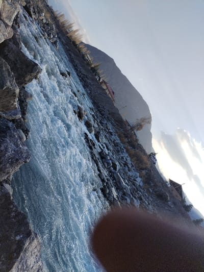 The image depicts a turbulent river flowing through a rocky, mountainous landscape in Qoraqiya, Uzbekistan. The river occupies the left and central portion of the frame, characterized by fast-flowing, light blue-green water with numerous white rapids. Its immediate left bank consists of large, dark grey boulders. The opposite bank, extending from the center-right towards the background, is a rocky, grey-brown slope with sparse, leafless deciduous vegetation.

Several man-made structures are visible along the right bank. A building with a prominent reddish-brown peaked roof and light-colored walls is situated further up the slope in the mid-distance. Another smaller, darker structure with a peaked roof, possibly incomplete, is located closer to the river on the same bank. A large, hazy grey mountain range dominates the background, rising sharply behind the river and buildings. The sky above is a clear, light blue with scattered white clouds, appearing brightest towards the upper right, indicating a low sun angle. A blurry, dark reddish-brown object, consistent with a finger or thumb, partially obstructs the lower-right corner of the image. No individuals or discernible actions are present within the scene. The environment is rugged, natural, and sparsely developed.