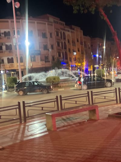 The image depicts an urban street scene at night in Beni Mellal, Morocco, focused on a traffic roundabout with a central fountain.
**Foreground:** The immediate foreground features a reddish-brown paver sidewalk. A dark-colored metal fence, consisting of vertical posts and horizontal bars, borders the sidewalk. A rectangular public bench, presenting a red top surface and a lighter base, is positioned on the sidewalk to the right.
**Midground:** A multi-lane roadway encircles a prominent, illuminated circular fountain. The fountain actively sprays multiple jets of water upwards and outwards. Small, trimmed green bushes line the perimeter of the fountain's base. Two dark-colored vehicles are in motion around the fountain; a dark SUV is on the left, and a dark sedan, potentially an extended model, with illuminated taillights, is on the right. The road surface reflects ambient light, suggesting a wet or highly reflective condition.
**Background:** A series of multi-story buildings, typically four to five floors high, forms the background. These structures exhibit light-colored facades consistent with local architecture, featuring arched windows and decorative elements. Ground floors house commercial businesses, identifiable by illuminated signage, including blue and yellow lights. Numerous bright streetlights and building lights illuminate the scene, creating reflections and a prominent vertical light flare. A tree with reddish-brown branches is partially visible in the upper right.
No individuals are distinctly visible within the frame. The scene conveys an active urban thoroughfare after dark.