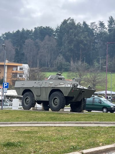 An olive green Portuguese Chaimite (V-200) armored personnel carrier is displayed as a monument on a raised platform composed of four light-colored concrete blocks. The vehicle is positioned centrally, facing slightly towards the right, and features large, rugged tires and a multi-faceted hull. A small plate on the front-right reads "MX-57-48". Chains are visible hanging from the front of the vehicle.

In the immediate foreground, a strip of green grass with sparse patches of brown extends across the bottom of the frame, separated by a concrete curb. Behind the armored vehicle, a paved road is visible, with a dark green minivan driving or parked on the right side. To the left of the armored vehicle, a blue "P" parking sign stands next to the road. Further left and behind the sign, a two-story building with a brown-orange section and a white section is partially visible, topped with orange roof tiles.

The background consists of a steeply rising hillside densely covered with trees. Closer to the midground, several bare deciduous trees are interspersed with dark green coniferous trees. Further up the slope, a dense forest of dark green conifers dominates. Multiple streetlights are scattered throughout the background. The sky above is uniformly overcast and light grey, indicating a cloudy day. No individuals are present in the scene. The location is the Rotunda da Chaimite in Lamego, Portugal.