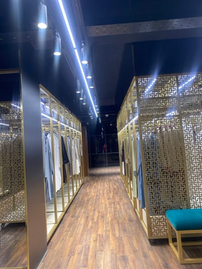 The image displays an interior view of a retail clothing establishment located in Narayanganj, Bangladesh. A central aisle, approximately 1.5-2 meters wide, extends into the background, flanked on both sides by gold-framed clothing display units. These units feature decorative, geometric lattice patterns in gold-colored metal and hold various long-sleeved garments, including kurtas or similar apparel, on hangers. Garment colors visible include white, grey, black, light blue, and beige. Some racks incorporate internal LED strip lighting.

The floor consists of dark brown wood-patterned laminate planks. The ceiling is dark, likely black, with exposed utility infrastructure, including cable trays. Overhead illumination is provided by a continuous central LED strip light and multiple cylindrical, downward-facing spot lights mounted along the ceiling.

At the distant end of the aisle, a large dark glass partition or window is present, displaying reflections and a very faint, indistinct reddish figure, possibly a person or mannequin, beyond it. In the foreground, on the right side of the aisle, a turquoise-upholstered bench with a gold metallic frame is situated adjacent to a clothing display unit. No individuals are clearly visible or actively engaged in the immediate foreground or midground.