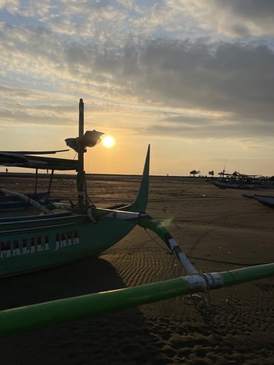 This image depicts a sunset or sunrise on a sandy beach in Negara, Indonesia. In the foreground and midground, a traditional Indonesian outrigger fishing boat, colored green with white and red accents, rests on the sand. The name "MALILI JAYA" is visible in white letters with red outlines on its hull. A mast-like structure rises from the boat, and a prominent green outrigger pole extends across the bottom of the frame, revealing worn white sections and coiled wires on the sand beneath.
The sun, a bright orange-yellow orb, is low in the sky, positioned to the right of the main boat's mast, casting long shadows. The sky is partially covered with scattered clouds, displaying warm hues near the horizon and lighter blue tones higher up. The sandy beach exhibits ripples and numerous footprints. In the midground and background, several silhouetted figures are walking or standing on the distant shore, and multiple other fishing boats are moored further along the beach. A cluster of small trees or bushes is visible on the horizon line to the right. Scattered debris, including what appears to be plastic refuse, is visible on the sand, particularly in the midground area.