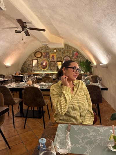 The image depicts the interior of a restaurant in Aix-en-Provence, France, characterized by a vaulted, off-white ceiling and a terracotta-tiled floor. A woman, with dark hair pulled back, wearing black-framed glasses and a light yellow vertical-striped button-up shirt, is seated at a wooden table in the foreground. She exhibits a subtle smile, looking toward the right out of frame, with her right hand raised, fingers touching her ear, and a ring visible on her right ring finger. On her table are a light blue placemat with a leaf pattern, two clear glasses (one containing water), a small blue-and-wood salt/pepper grinder, a knife, and a partially visible glass with ice and lime.
The restaurant is furnished with multiple rectangular dark wooden tables and dark brown upholstered chairs; all tables are set with similar placemats, white napkins, and various types of glassware, indicating readiness for service. The far wall features a textured, dark patterned wallpaper serving as a backdrop for a densely arranged collection of framed artwork and mirrors of assorted sizes and shapes. Overhead, a brass and dark wood ceiling fan with light fixtures is mounted. The scene is softly illuminated by wall-mounted uplights highlighting the ceiling arches. A large green leafy plant is visible among the background tables.