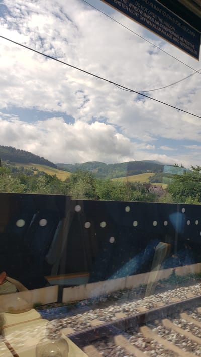The image shows a view from a moving train, likely in a rural area. In the foreground, there are train tracks and a dark barrier with circular reflections. Beyond the barrier, a lush green landscape with rolling hills and scattered trees is visible. Some houses are nestled among the trees on a hillside. The sky is partly cloudy with bright sunlight. A sign above the window reads, "DO NOT OPEN THE WINDOW, THE WINDOW MAKES AIR-CONDITIONING INEFFECTIVE. ENSTER NICHT OFFNEM, KLIMAANLAGE WIRD WIRKUNGSLOS." The text suggests the train is equipped with air conditioning and opening the window would disrupt its effectiveness. Based on the language on the sign and the general scenery, the location is likely in a Central European country. Although specific details to pinpoint Maršová-Rašov, Slovakia are not definitively present, the landscape is consistent with rural areas in Slovakia.