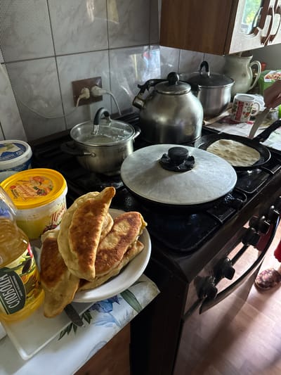 This image depicts a close-up view of a gas stove in a kitchen, likely in Valday, Russia. Several cooking items are present. On the stovetop, a metal teapot, a covered pot, and two pans are visible. One pan contains a flatbread, and another is covered with a large metal lid. A plate of fried pastries, possibly chebureki or pirozhki, is situated on the left side of the stove. Next to the plate, a bottle of sunflower oil with a label in Russian is placed on a white tray. Two tubs of dairy products are also on the counter to the left. The kitchen backsplash is tiled with a light-colored pattern. In the background, a wooden cabinet is visible. A person's hand is seen in the upper right corner, holding a knife, suggesting activity in the kitchen. The floor appears to be light-colored wood.
