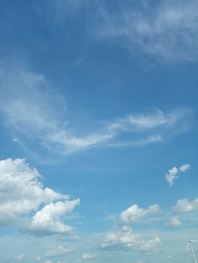 A sky filled with blue and white clouds is visible in the image. The clouds are scattered across the sky, with some appearing wispy and others more dense and fluffy. A portion of a white wind turbine is visible on the right side of the image, suggesting the location is an open area possibly used for renewable energy generation. The scene depicts a clear day with a vast expanse of sky.