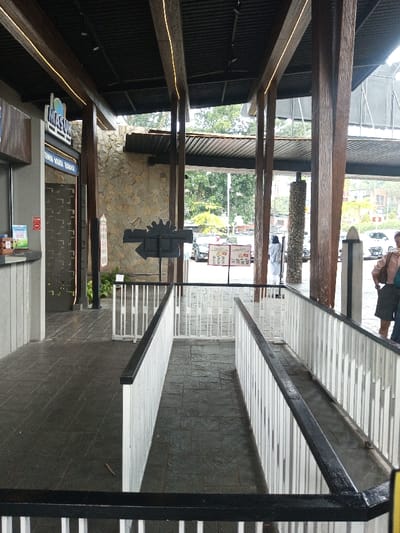 The image depicts a semi-outdoor commercial establishment located in Tân Kỳ Commune, Vietnam. The scene features a long, narrow pathway defined by white vertically slatted wooden railings with black top rails, leading away from the viewer towards a brighter outdoor area. The path's dark grey stamped concrete flooring suggests a slight downward slope.
On the left, a grey service counter is visible, topped with small signs including a red 'No Smoking' emblem and various leaflets. Above this counter, a prominent light blue sign displays "MASJUR" in white text with a logo, and "DUNGAN WARA, BERSAUA" below. Adjacent to the counter is an entrance with a patterned, dark grey door. The left side of the structure features a textured wall of light brown and beige stones.
The overhead structure consists of a dark, corrugated metal roof supported by numerous dark wooden beams. Yellow LED strip lighting runs along the undersides of some beams, illuminating the covered area. Several rough-hewn dark wooden pillars and one cylindrical stone-clad pillar support the roof.
In the mid-ground, a dark, arrow-shaped sign with a sun-like or cog-like motif points left. Beyond the railings and pillars, the background reveals an outdoor setting with green trees and multiple vehicles, including white cars and a darker SUV, parked on what appears to be a road or parking lot. Several vertical promotional signs with images and text are visible near the vehicles.
Two individuals are partially visible in the background on the right: one person wearing a long white garment and a head covering stands near a stone pillar, and closer to the foreground, a second person wearing a light brown top and a dark skirt/shorts stands with their back mostly to the camera. Another person is partially obscured behind them. The environment is naturally lit from the open-air design.