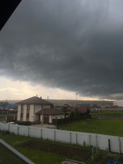 The image depicts a cloudy sky over a residential area, possibly the city of Solontsy, Russia. In the foreground, a white corrugated metal fence runs horizontally, partially obscuring a grassy yard with some tilled soil. Beyond the fence, a two-story house with a light-colored facade and a dark brown roof is visible. Adjacent to this house is a garage with a brown door. Several other houses with different colored roofs, including blue and brown, are scattered throughout the scene, along with more fences and some trees. In the background, industrial buildings and a construction crane suggest the presence of ongoing development or industrial activity. The sky is dominated by heavy, dark grey storm clouds, with a faint glow of light breaking through on the horizon, indicating a potential storm or overcast weather. No people are visible in the image.