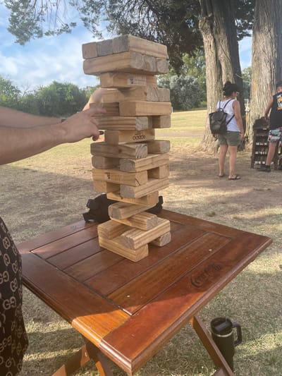 A Jenga tower sits on a wooden Coca-Cola branded table outdoors. Two hands are attempting to remove a piece. In the background, a person in a white shirt and green shorts walks by, a black backpack over their shoulder. Further back, there is a bean bag toss game and trees. The ground is covered in grass and dirt.
