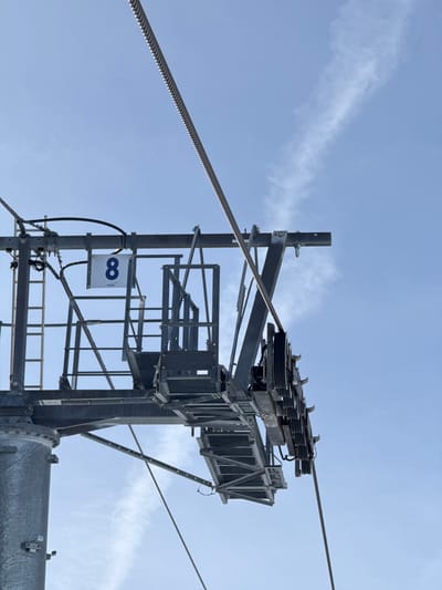 The image displays the upper section of a metallic ski lift pylon, identified as Pylon 8 of a POMA-manufactured system, located in La Salle-les-Alpes, France. The pylon's steel framework, comprising horizontal and diagonal beams, supports an integrated maintenance platform and ladder sections. A sheave assembly, consisting of multiple dark rollers, is visible on the right side of the structure. A thick, ridged steel haul rope runs diagonally from the upper left to the lower right, passing over the sheave assembly. A white rectangular sign bearing the black numeral "8" and the text "POMA" is affixed to the pylon's upper structure. Minor electrical conduits or sensor wires are visible along the pylon's components. The background is a clear, bright blue sky with a faint, elongated white contrail or wispy cloud stretching diagonally across the upper right quadrant. No individuals, animals, or specific actions are depicted.