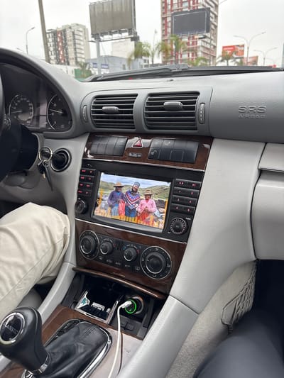 The image presents a detailed interior view from the front passenger seat of a vehicle, looking forward through the windshield into an urban environment identified as La Victoria, Peru.
Inside the vehicle, a light grey dashboard with a textured finish dominates. On the driver's side (left), a partial view of the steering wheel and instrument cluster is visible, showing a speedometer marked up to 260 and a tachometer up to 7,000 RPM. Keys hang from the ignition switch below the instrument cluster. The driver's left arm and a tan pant leg are visible on the lower left. The central console features a dark wood-grain trim. Above the infotainment system, a row of buttons includes the hazard lights, seat heating controls, and central locking. The infotainment screen displays a colorful image of three individuals in traditional Andean attire (hats, patterned ponchos/shawls) standing outdoors in a hilly, grassy landscape. Flanking the screen are various control buttons labeled MUTE, BAND, AMS, NAVI, MEDIA, BT, VOL, TUNE, EQ, and numbered presets from 1 to 6. Below the screen, the climate control panel features three rotary knobs for temperature and fan speed, with an "AUTO AC" button. Further down, the gear selector for an automatic transmission is visible, topped with a black knob bearing the word "CLASSIC." Adjacent to the gear selector, a small compartment houses a car charger adapter with a glowing green LED, connected to a white USB cable. On the far right, a light grey mesh storage net is affixed to the passenger-side footwell. A leg clad in dark pants is partially visible in the lower right, indicating a passenger.
Through the windshield, a cloudy, overcast sky hangs over a cityscape. Multiple multi-story buildings of varying designs are visible, including residential apartments and commercial structures. A prominent red-colored building stands in the middle distance. Several large billboards are present, one appearing to be blank or unlit, and another on the right displaying what appears to be a food advertisement. Streetlights with multiple arms and overhead wires are visible. Palm trees dot the urban landscape, suggesting a warm climate. The overall scene depicts a typical busy urban setting under a dull, daytime sky.