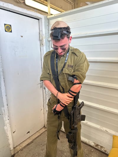 A young man, wearing a kippah and a dark green military uniform with the word "צה"l" (Tzahal) on the right sleeve, is standing in a hallway. He is wearing black tefillin on his left arm and is holding a black rifle with his right hand. His head is bowed and his eyes are closed. He is wearing sunglasses on top of his head. The hallway is painted white with horizontal lines and has a white door at the left of the frame. The floor is concrete and there is a white floorboard leading out of the room on the left side. The image was taken in the city of Mazkeret Batya, Israel, at the location of coordinates: latitude: 31.833, longitude: 34.82.