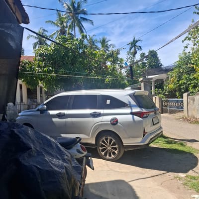 The image depicts a white SUV parked on a dirt road in front of a house in Reudeuep, Indonesia. The car is positioned on the right side of the frame, with its rear end facing towards a blue and white gate. A black motorcycle, partially covered by a dark tarp, is situated to the left of the SUV. In the background, a house with a red-tiled roof and wooden stilts is visible, surrounded by lush green trees and palm trees. The sky is clear blue with a few wispy clouds. The scene appears to be a typical residential area in a tropical climate.