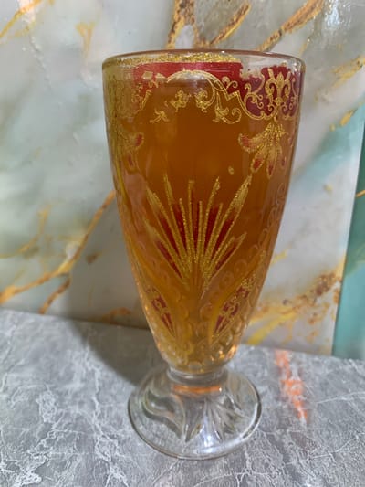 A glass filled with amber liquid stands on a marble-patterned surface. The glass is decorated with red and gold intricate patterns. The background features a marbled wall with gold veining. The scene appears to be an indoor setting, possibly a table in a home or establishment, in the city of Yuada-Gulrypsh, Abkhazia. There are no people or actions depicted in the image.
