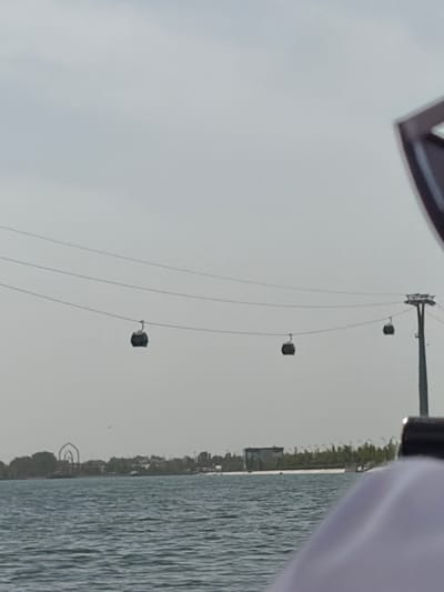 The image presents an expansive landscape captured from a low vantage point, likely from a boat or lakeside. The dominant feature is a cableway system stretching across a wide body of water under an overcast sky.
In the immediate foreground, at the top right of the frame (when viewed correctly oriented), the partial outline of a dark, angular object is visible, possibly part of a boat or a viewing platform. Adjacent to it, a soft, light-colored fabric, resembling clothing, is also partially seen.
The main focus of the scene is a series of taut cables suspended over the water. A tall, dark pylon or tower stands on the far bank, anchoring one end of the cable system. Several dark, rectangular cabins are suspended from these cables at regular intervals, suggesting a functioning cable car or ski lift type of transportation. The sky is a uniform, pale grey, indicating an overcast day.
Below the cable system, the surface of the large water body stretches across the midground, displaying subtle ripples. The far bank, beyond the water, features a narrow strip of land covered with green vegetation and some indistinct structures, possibly buildings, hinting at human presence.
The scene depicts a calm, daytime environment with no apparent human activity in progress apart from the infrastructure itself. The overall impression is one of tranquility, with man-made structures integrated into the natural landscape. Given the context of Kuchtumgali, Uzbekistan, this vista likely showcases the Charvak Reservoir area, known for its scenic views and recreational facilities, including cableways. No visible text is present in the image.